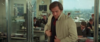 Movie still from “A Touch of Class” (1973), directed by Melvin Frank – A man wearing a trench coat and tie in an office setting; Medium shot, Over the shoulder angle