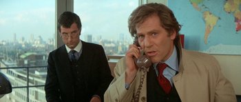 Movie still from “A Touch of Class” (1973), directed by Melvin Frank – A man on the phone with another man in the background; Medium shot, Over the shoulder angle