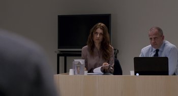 Movie still from “A War” (2015), directed by Tobias Lindholm – A woman sitting in front of a tv in front of a wooden table; Medium shot, Over the shoulder angle