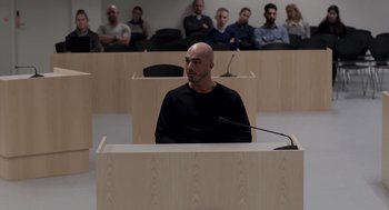 Movie still from “A War” (2015), directed by Tobias Lindholm – A man in a black shirt is in a court room; Wide shot, Over the shoulder angle