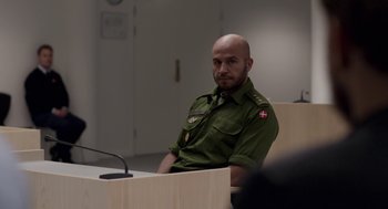 Movie still from “A War” (2015), directed by Tobias Lindholm – A man sitting in front of a microphone in front of a podium; Medium shot, Over the shoulder angle