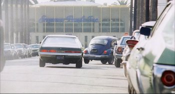 Movie still from “A Woman Under the Influence” (1974), directed by John Cassavetes – A group of cars driving down a street next to a building; Wide shot, Low angle