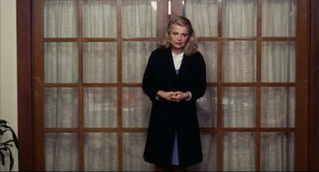 Movie still from “A Woman Under the Influence” (1974), directed by John Cassavetes – A woman standing in front of a window wearing a black coat; Medium shot, Low angle