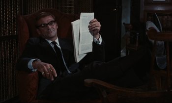 Movie still from “Accident” (1967), directed by Joseph Losey – A man sitting in a chair holding a newspaper; Medium shot, Low angle