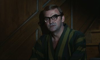 Movie still from “Accident” (1967), directed by Joseph Losey – A man wearing a robe and glasses in a dark room; Close Up shot, Low angle