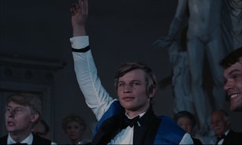Movie still from “Accident” (1967), directed by Joseph Losey – A man raising his hand in the air in front of a group of people; Medium shot, Low angle