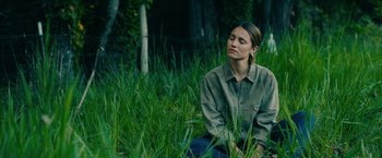 Movie still from “Acidman” (2022), directed by Alex Lehmann – A woman sitting in a field of tall green grass; Medium shot, Low angle
