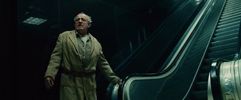 Movie still from “Across the Universe” (2007), directed by Julie Taymor – An old man in a trench coat standing next to an escalator; Medium shot, Low angle