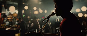 Movie still from “Across the Universe” (2007), directed by Julie Taymor – A man is speaking into a microphone at a concert; Medium shot, Over the shoulder angle