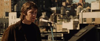 Movie still from “Across the Universe” (2007), directed by Julie Taymor – A man standing in front of a microphone on top of a building; Close Up shot, Over the shoulder angle