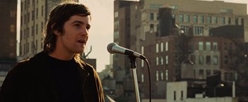 Movie still from “Across the Universe” (2007), directed by Julie Taymor – A man standing in front of a microphone on top of a building; Close Up shot, Over the shoulder angle