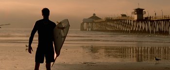 Movie still from “Act of Valor” (2012), directed by Scott Waugh – A man holding a surfboard standing on the beach at sunset; Wide shot, Low angle