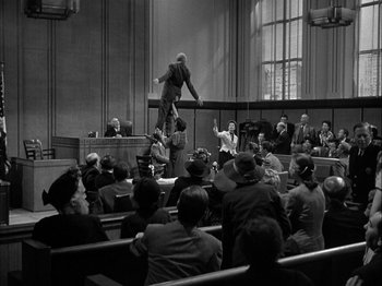 Movie still from “Adam's Rib” (1949), directed by George Cukor – A man standing on the back of a judge's chair in front of a group of people; Wide shot, Low angle