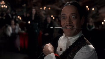 Movie still from “Addams Family Values” (1993), directed by Barry Sonnenfeld – A man holding a fork in his mouth while standing in front of a crowd of people; Close Up shot, Low angle