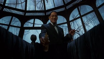 Movie still from “Addams Family Values” (1993), directed by Barry Sonnenfeld – A man holding a bottle and smoking a cigarette in front of a window; Medium shot, Low angle