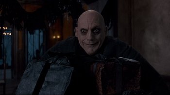 Movie still from “Addams Family Values” (1993), directed by Barry Sonnenfeld – A man with a bald head is holding some suitcases; Close Up shot, Over the shoulder angle
