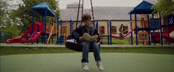 Movie still from “Admission” (2013), directed by Paul Weitz – A young boy sitting on a swing reading a book; Wide shot, High angle