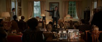 Movie still from “Admission” (2013), directed by Paul Weitz – A man standing in front of a group of people in a living room; Wide shot, Over the shoulder angle