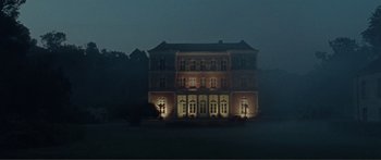 Movie still from “Adoration” (2019), directed by Fabrice du Welz – A large building lit up at night in the fog; Extreme Wide shot, Low angle