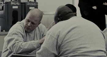 Movie still from “Adulthood” (2008), directed by Noel Clarke – A man sitting next to another man in a room; Medium shot, Over the shoulder angle