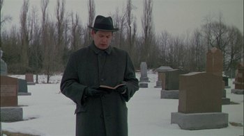 Movie still from “Affliction” (1997), directed by Paul Schrader – A man in a hat and coat standing in the snow reading a book in front of a cemetery; Medium shot, Low angle
