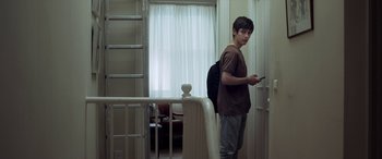 Movie still from “After Love” (2020), directed by Aleem Khan – A young man standing in front of a door holding a cell phone; Medium shot, Low angle