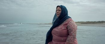 Movie still from “After Love” (2020), directed by Aleem Khan – A woman in a pink dress and a blue head scarf standing in front of a body of water; Medium shot, Low angle