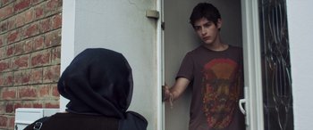 Movie still from “After Love” (2020), directed by Aleem Khan – A young man holding a door handle in front of a door; Medium shot, Over the shoulder angle