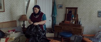 Movie still from “After Love” (2020), directed by Aleem Khan – A woman holding a bag of garbage in a living room; Medium shot, Over the shoulder angle