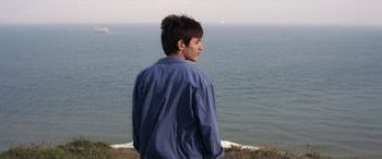 Movie still from “After Love” (2020), directed by Aleem Khan – A man standing on top of a hill looking out at the ocean; Medium shot, High angle