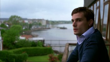 Movie still from “After the Wedding” (2006), directed by Susanne Bier – A man sitting on top of a wooden railing near a body of water; Close Up shot, Over the shoulder angle