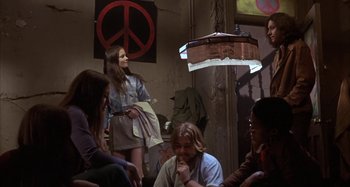 Movie still from “Alice's Restaurant” (1969), directed by Arthur Penn – A group of people sitting in a room with a peace sign on the wall behind them; Wide shot, High angle