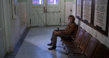 Movie still from “Alice's Restaurant” (1969), directed by Arthur Penn – A man sitting on a bench in front of a door way; Wide shot, High angle