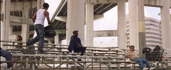 Movie still from “All About the Benjamins” (2002), directed by Kevin Bray – A man sitting on top of a bench near a bridge; Wide shot, Low angle