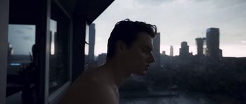 A shirtless man looking out of a window at a city skyline