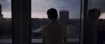 A shirtless man looking out of a window at a city