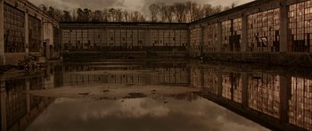 Movie still from “All the Real Girls” (2003), directed by David Gordon Green – A reflection of a building in a puddle of water near a forest; Extreme Wide shot, Low angle