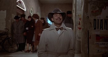 Movie still from “Amarcord” (1973), directed by Federico Fellini – A man in a trench coat and a bow tie standing in a hallway; Medium shot, Over the shoulder angle