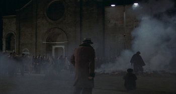 Movie still from “Amarcord” (1973), directed by Federico Fellini – A group of people standing in front of a building with smoke coming out of it; Extreme Wide shot, High angle