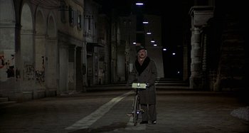 Movie still from “Amarcord” (1973), directed by Federico Fellini – An old man standing next to a bicycle in the middle of a street; Wide shot, High angle