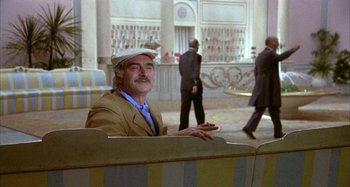 Movie still from “Amarcord” (1973), directed by Federico Fellini – An older man sitting on top of a bench in front of a building; Medium shot, Over the shoulder angle