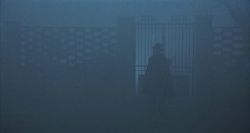 Movie still from “Amarcord” (1973), directed by Federico Fellini – A man in a black coat and a black hat is standing in the fog; Wide shot, Over the shoulder angle