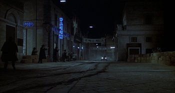 Movie still from “Amarcord” (1973), directed by Federico Fellini – A street at night with people standing on the sidewalk and cars driving down the street; Wide shot, High angle