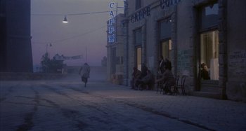 Movie still from “Amarcord” (1973), directed by Federico Fellini – A group of people sitting on a sidewalk near a building; Extreme Wide shot, High angle