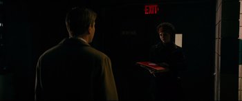 Movie still from “American Pastoral” (2016), directed by Ewan McGregor – A man holding a red box in front of another man in front of an exit sign; Medium shot, Over the shoulder angle