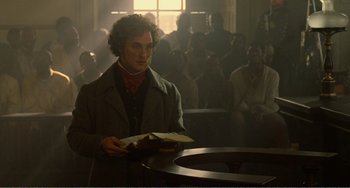 Movie still from “Amistad” (1997), directed by Steven Spielberg – A man holding a book while standing in front of a group of people; Medium shot, Over the shoulder angle