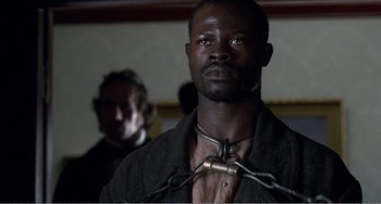 Movie still from “Amistad” (1997), directed by Steven Spielberg – A man with a chain around his neck and another man in the background; Close Up shot, Low angle