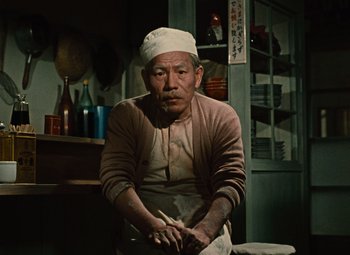 An older man wearing a white hat and a brown apron