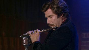 Movie still from “Anchorman: The Legend of Ron Burgundy” (2004), directed by Adam McKay – A man is playing the flute while sitting at a microphone; Close Up shot, Low angle