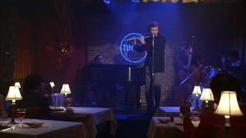 Movie still from “Anchorman: The Legend of Ron Burgundy” (2004), directed by Adam McKay – A man holding a microphone while standing in front of a piano; Wide shot, High angle